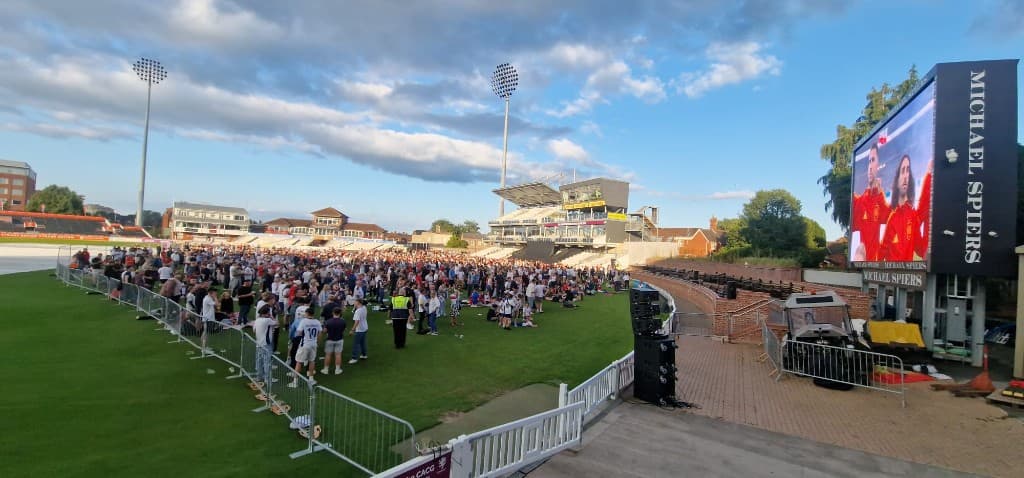A crowd on the outfield at a cricket ground, with a big screen beside the stand